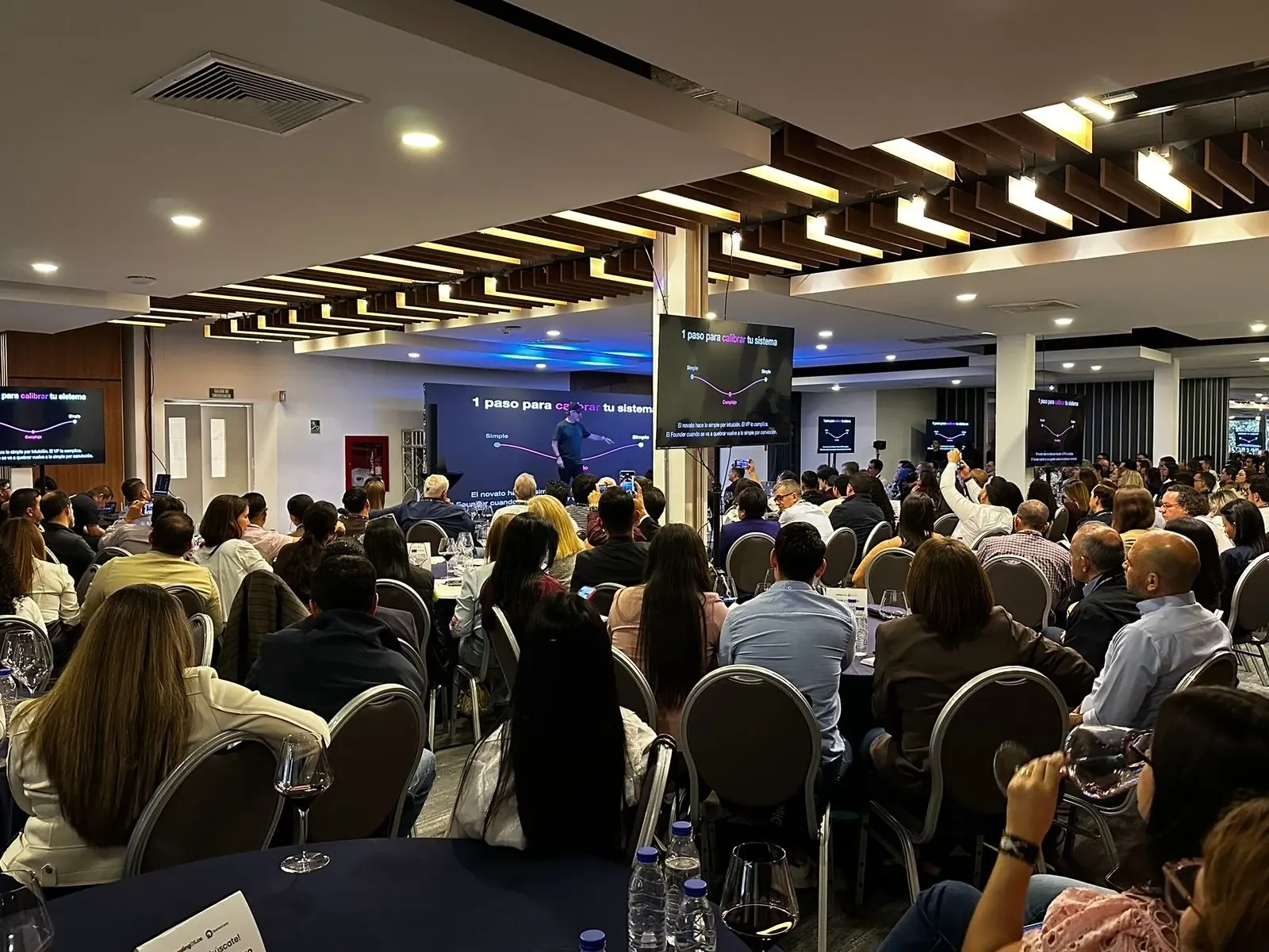 Sala llena en el workshop de San Cristóbal, con la diapositiva «1 paso para calibrar tu sistema» en todas las pantallas